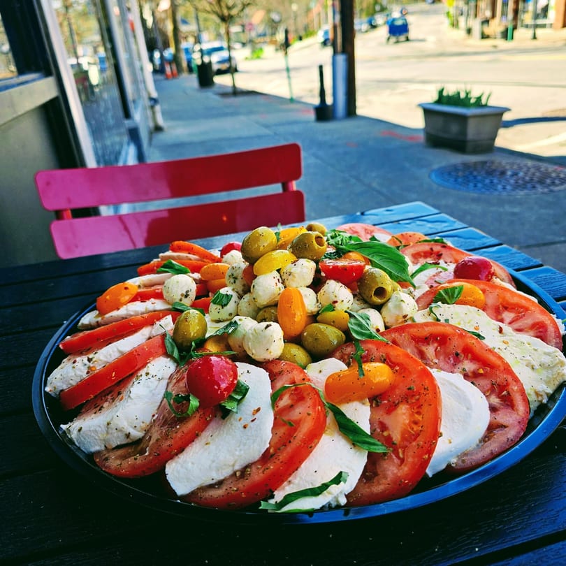Caprese salad with sliced tomatoes, mozzarella, basil, olives, and cherry tomatoes on a black platter.