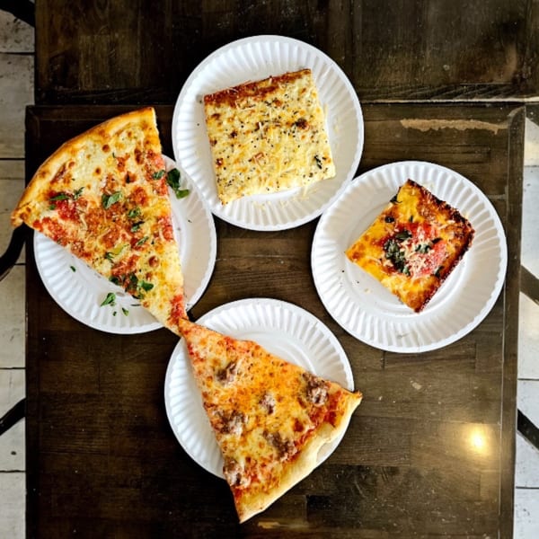 Four slices of pizza, including plain, Sicilian, and sausage, on paper plates on a wooden table.