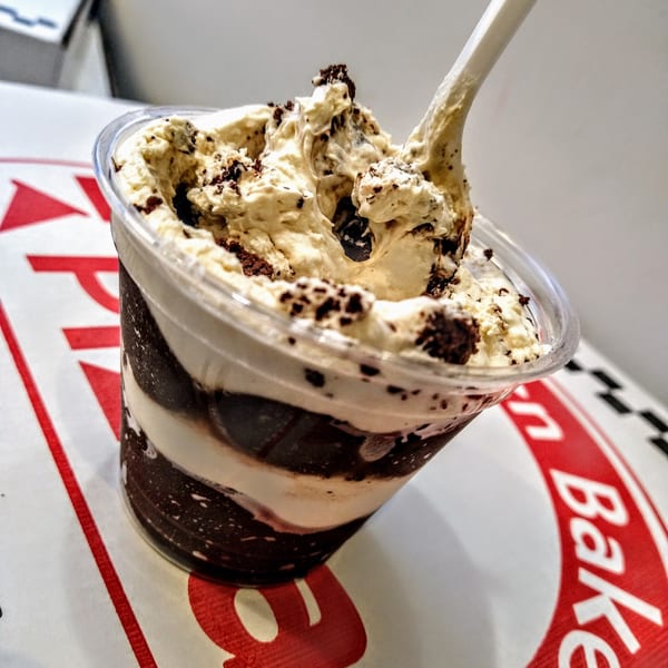 A plastic cup of layered chocolate and vanilla ice cream with cookie crumbles and a spoon.