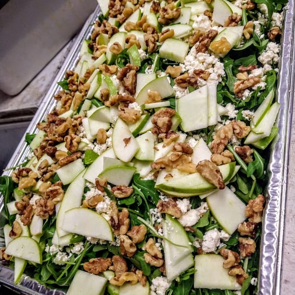 Arugula salad with green apple slices, walnuts, and crumbled feta cheese in a metal tray.