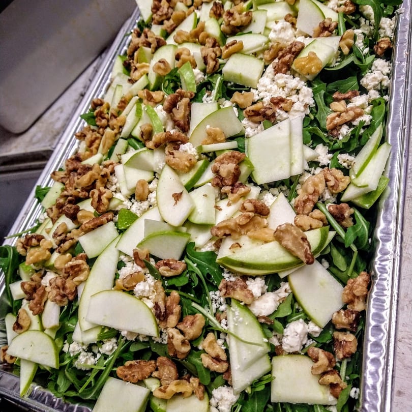 Arugula salad with green apple slices, walnuts, and crumbled feta cheese in a metal tray.