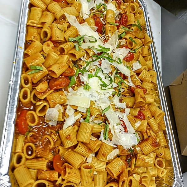 A large catering tray of rigatoni pasta with tomato sauce, cherry tomatoes, basil, and parmesan cheese.