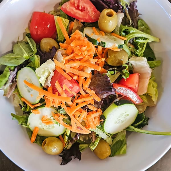 Fresh garden salad with mixed greens, tomatoes, cucumbers, olives, and shredded carrots in a white bowl.