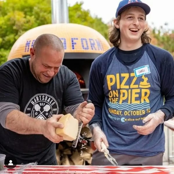 Two men, one grating cheese, the other smiling, in front of a pizza oven.