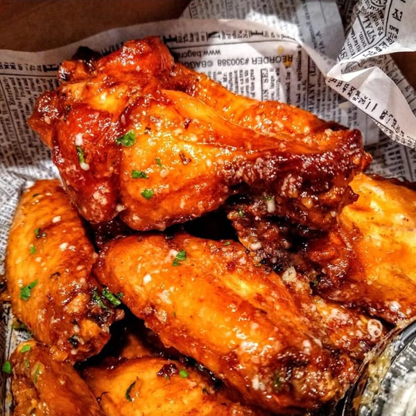 Close-up of glossy, sauced chicken wings with herbs and cheese, served in a paper-lined container.