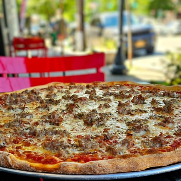 A large, freshly baked sausage pizza from Hartsdale House of Pizza, with a blurred outdoor background.